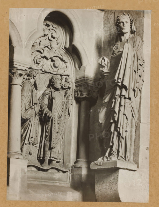 Sculpture in Bamberg Cathedral, album page, photograph, This photo is part of an album., number: '175', Bamberg, 1900 - c. 1920, photographer, Bavaria, paper, baryta paper, gelatin silver print, developed gelatin silver print, height 226 mm x width 98 mm, church (exterior), Bamberg Cathedral, A grayscale photograph showing a close view of carved stone figures and architectural details mounted on a rectangular background. On the right a tall draped statue stands on a projecting plinth, wearing a long flowing garment with deep folds and holding a wrapped object near the chest; the figure's face is turned slightly upward and is covered at the top by a head cloth. To the left of this figure is a column with a molded capital supporting an arch; beneath the arch are two smaller carved figures in ornate robes, one wearing a crown and the other with a hooded mantle, both standing on a decorated base and flanked by carved foliage and scrollwork. Above the smaller figures an arched niche frames ...