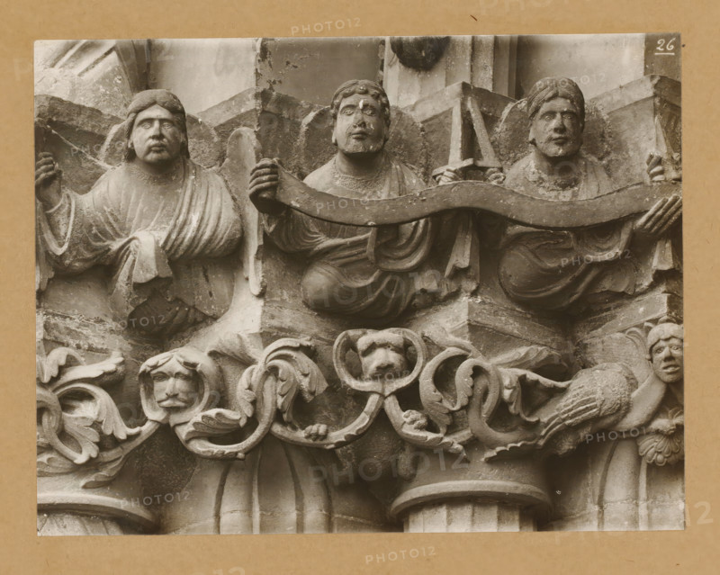 Marienpforte, left. Chapter zone middle from north-east., Detail of a capital of the Gnadenpforte in Bamberg Cathedral, album page, photograph, This photo is part of an album., number: '26.', Bamberg, 1900 - c. 1920, photographer, Bavaria, paper, baryta paper, gelatin silver print, developed gelatin silver print, height 169 mm x width 220 mm, church (exterior), Bamberg Cathedral, A stone relief showing three robed figures in a row holding a curved band, each with distinct facial features and hairstyles, a central figure with a beard and two flanking figures with shorter hair; below them an ornate carved frieze with foliate scrolls, a small human head with stylized hair and a central carved animal face surrounded by curved decorative elements, and a partially visible kneeling figure on the right holding part of the scrollwork; the stone surface shows weathering and small chips, and the composition is set against a plain rectangular backing.