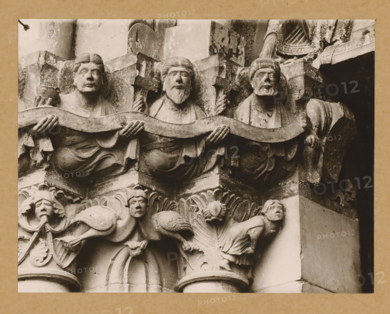 Marienpforte, left. Capital zone Right front, Detail of a capital of the Gnadenpforte in Bamberg Cathedral, album page, photograph, This photo is part of an album., number: '27.', Bamberg, 1900 - c. 1920, photographer, Bavaria, paper, baryta paper, gelatin silver print, developed gelatin silver print, height 168 mm x width 219 mm, church (exterior), Bamberg Cathedral, Stone sculpted figures arranged horizontally, each with distinct facial features and hair, holding a wavy band across their chests; they wear draped garments and one holds a small vessel while others grip carved crosses. Below them are ornate carved elements including leafy motifs and two smaller humanlike figures positioned on their hands and knees with expressive faces and detailed hair. The stone shows weathering and textured surfaces with areas of darker staining and shadowed recesses, and parts of the surrounding architectural carving and columns are visible at the sides.