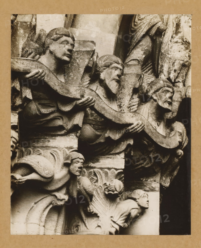 Detail of a capital of the Gnadenpforte in Bamberg Cathedral, Marienpforte, left. Capital zone Right front, album page, photograph, This photo is part of an album., number: '28.', Bamberg, 1900 - c. 1920, photographer, Bavaria, paper, baryta paper, gelatin silver print, developed gelatin silver print, height 205 mm x width 168 mm, church (exterior), Bamberg Cathedral, A close-up photograph of a carved stone relief mounted on a tan mat, showing multiple human figures in varying poses. The upper portion features three older-looking figures with carved hair and beards, two facing sideways and one turned toward the right, each holding or supporting a long, curved stone element that weaves across the composition. Below them, a crouching figure with a rounded face and stylized hair peers outward, positioned among carved architectural elements and ornamental foliage. The stone surfaces show weathering and textured patina, with deep shadows highlighting folds in drapery, carved facial features...