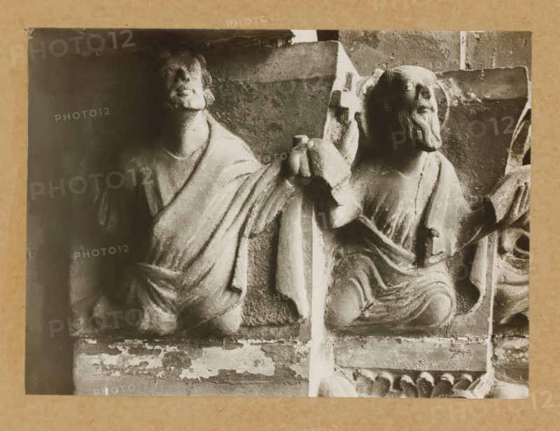 Detail of a capital of the Gnadenpforte in Bamberg Cathedral, Marienpforte, r. Kapitellzone, li vS, album page, photograph, This photo is part of an album., number: '29.', Bamberg, 1900 - c. 1920, photographer, Bavaria, paper, baryta paper, gelatin silver print, developed gelatin silver print, height 162 mm x width 221 mm, church (exterior), Bamberg Cathedral, A cropped black-and-white photograph of two carved stone figures seated side by side, each wearing draped garments with visible folds; the figure on the left has a rounded face and short hair, the figure on the right has a beard and a halo-like circular carving behind the head; both figures rest on a textured stone background with a vertical divider between them and a horizontal ledge below that shows chipped and weathered surfaces.