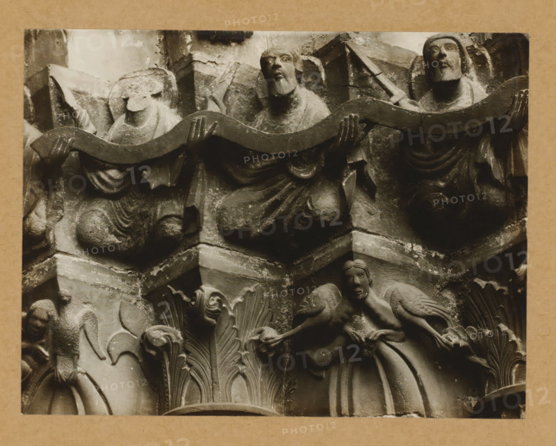 Detail of a capital of the Gnadenpforte in Bamberg Cathedral, Marienpforte, right-hand chapter zone, center of south-east, album page, photograph, This photo is part of an album., number: '32.', Bamberg, 1900 - c. 1920, photographer, Bavaria, paper, baryta paper, gelatin silver print, developed gelatin silver print, height 167 mm x width 219 mm, church (exterior), Bamberg Cathedral, A close-up of a carved stone relief showing several human figures and decorative foliage elements. Three robed figures in the upper portion hold a long, undulating band, with two of them kneeling and one more upright, their faces showing carved features and stylized beards. Below them, ornate acanthus-like leaves form a central vertical motif flanked by smaller carved figures; one figure on the right appears to be seated with a hand touching its face and another figure on the left has a crouched posture. The stone shows weathering and surface texture, with visible joints and layered architectural elements a...