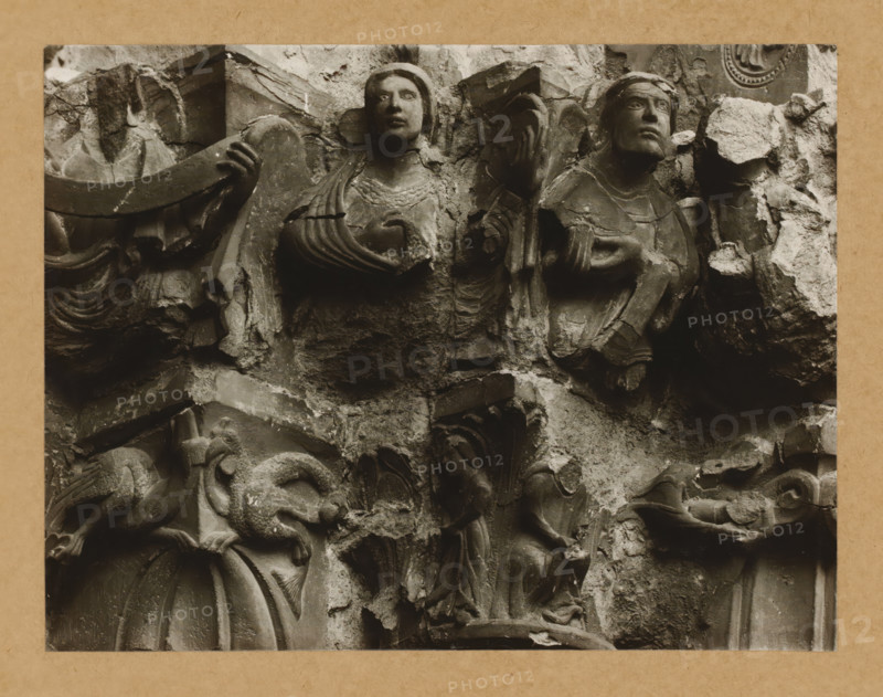 Detail of a capital of the Gnadenpforte in Bamberg Cathedral, Marienpforte, r. Kapitellzone, rv SO., album page, photograph, This photo is part of an album., number: '33.', Bamberg, 1900 - c. 1920, photographer, Bavaria, paper, baryta paper, gelatin silver print, developed gelatin silver print, height 168 mm x width 220 mm, church (exterior), Bamberg Cathedral, A close-up view of a weathered stone relief shows several carved human figures and decorative elements arranged on a rough background. Two larger upper figures face forward with detailed facial features, draped garments, and stylized wings or flowing cloth around their shoulders, one with a hand raised near the chest and the other with a hand extended to the side. Below and around them are smaller, partially damaged figures and ornamental shapes, including curved drapery, fragmented architectural details, and rough, chipped stone surfaces. The entire scene is monochromatic with strong contrasts between smooth carved areas and co...