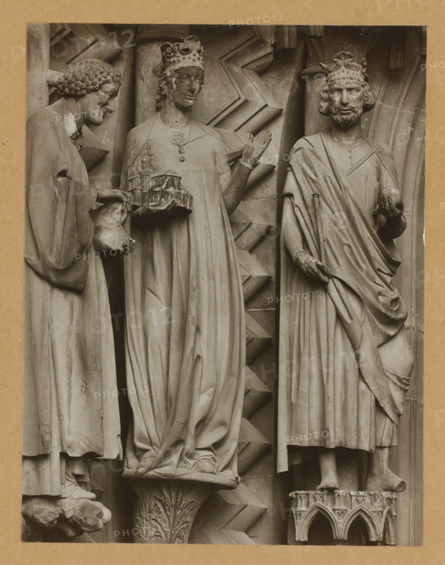 Adamspforte, left wall of the North., Detail of the Adamspforte in Bamberg Cathedral, album page, photograph, This photo is part of an album., number: '36.', Bamberg, 1900 - c. 1920, photographer, Bavaria, paper, baryta paper, gelatin silver print, developed gelatin silver print, height 210 mm x width 163 mm, church (exterior), Bamberg Cathedral, Three stone figures carved in relief stand side by side against a textured architectural background, each wearing long draped garments and ornate crowns; the left figure leans slightly forward with curly hair and extends hands holding a small sculpted object, the central figure faces forward with one hand raised and wears a necklace while holding a model-like structure, and the right figure has a beard and holds a spherical object in one hand with the other hand resting on drapery, all figures show detailed folds in their robes and stand on carved pedestals with geometric and floral motifs.