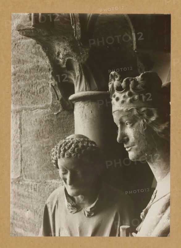 Adamspforte, Stephen and Kunigunde (Heads), Detail of the Adamspforte in Bamberg Cathedral, album page, photograph, This photo is part of an album., number: '38.', Bamberg, 1900 - c. 1920, photographer, Bavaria, paper, baryta paper, gelatin silver print, height 223 mm x width 161 mm, Person, Stephen (martyr), church (exterior), Bamberg Cathedral, A close-up monochrome photograph of two stone sculptures positioned side by side against a textured stone wall and a dark background, showing the right-profile of a crowned figure with carved hair, a serene facial expression, and a detailed ornate crown, next to a lower-headed figure with tightly curled hair, downcast eyes, and a simple draped garment, with visible weathering and surface texture on both sculptures and architectural stone elements above and behind them.