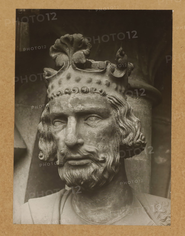 Adamspforte (Kaiser Heinrich) (Head from front), Detail of the Adamspforte in Bamberg Cathedral, album page, photograph, This photo is part of an album., number: '45.', Bamberg, 1900 - c. 1920, photographer, Bavaria, paper, baryta paper, gelatin silver print, gelatin silver developing print, height 224 mm x width 163 mm, Person, Saint Henry II (Holy Roman Emperor), church (exterior), Bamberg Cathedral, A close-up photograph of a stone sculpture showing the head and upper torso of a bearded man wearing a detailed crown with decorative leaf-like and curled elements, visible weathering and surface texture on the stone, wavy hair framing the face, carved eyes and a solemn expression, a portion of draped clothing across the shoulders, and a dark background with a cylindrical column partially visible behind the head.