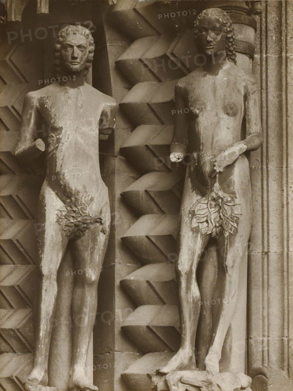 Detail of the Adamspforte in Bamberg Cathedral, Adamspforte, Adam and Eve in front, album page, photograph, This photo is part of an album., number: '49.', Bamberg, 1900 - c. 1920, photographer, Bavaria, paper, baryta paper, gelatin silver print, height 213 mm x width 161 mm, church (exterior), Bamberg Cathedral, A sepia-toned photograph of two stone statues of nude human figures mounted side by side against a carved architectural backdrop, both figures showing weathered surfaces and missing or damaged hands, the left figure with short curly hair and a solemn facial expression and the right figure with longer wavy hair and a faint smile, each figure having sculpted leaf coverings at the groin, the backdrop featuring a repeating geometric zigzag pattern between the figures and fluted stonework at the right edge, the lower portion showing the figures' legs and feet with a worn pedestal beneath the right foot and the entire image framed by a lighter border.