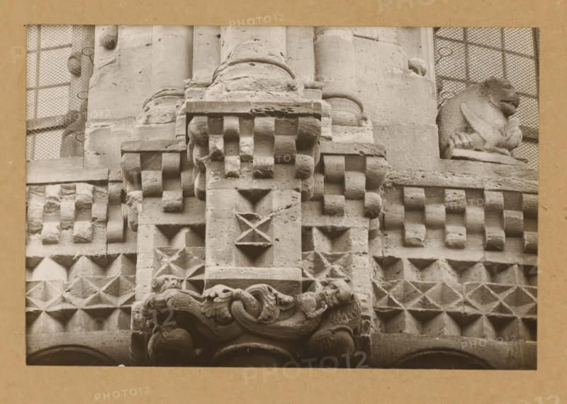 East Church, 1. Frieze 3., View of the sculpture of a frieze of Bamberg Cathedral, album page, photograph, This photo is part of an album., number: '5.', Bamberg, 1900 - c. 1920, photographer, Bavaria, paper, baryta paper, gelatin silver print, height 141 mm x width 219 mm, church (exterior), Bamberg Cathedral, A close-up of a carved stone façade showing geometric protruding blocks and pyramidal shapes, a central vertical stone column with sculpted decorative elements including rounded blocks and recessed diamond and triangular recesses, ornate carved scrollwork and foliage near the bottom, a sculpted animal figure perched on a corner ledge on the right, and mesh-covered window openings visible in the upper corners.