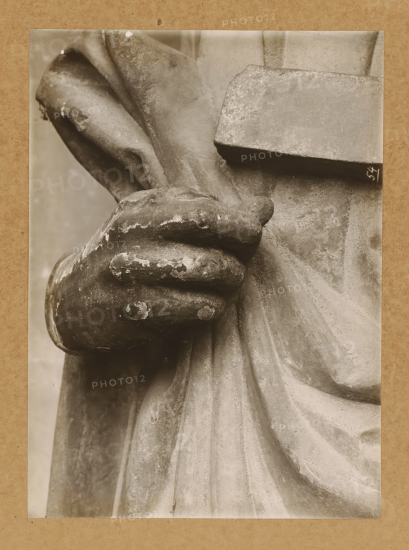 Adamspforte, r. Hand, Detail of the Adamspforte in Bamberg Cathedral, album page, photograph, This photo is part of an album., number: '54', Bamberg, 1900 - c. 1920, photographer, Bavaria, paper, baryta paper, gelatin silver print, developed gelatin silver print, height 165 mm x width 227 mm, church (exterior), Bamberg Cathedral, A close-up sepia-toned photograph of a weathered stone hand clutching a rectangular slab, showing chipped and flaking surface texture on the fingers and knuckles, a draped garment with deep folds and soft shadows, and the rough, pitted surface of the slab and surrounding stone.