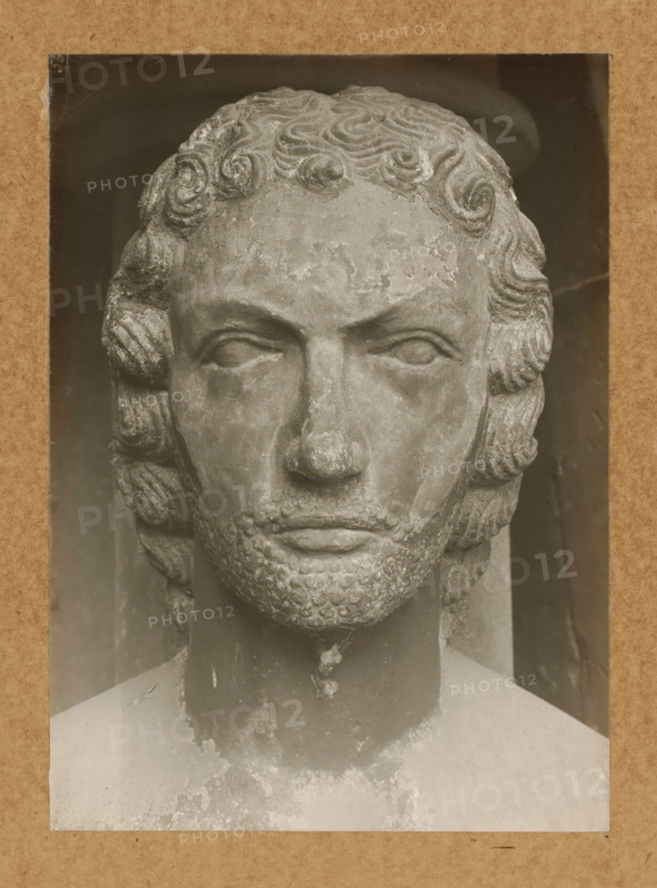 Adamspforte, Adam, Head from front, Detail of the Adamspforte in Bamberg Cathedral, album page, photograph, This photo is part of an album., number: '56', Bamberg, 1900 - c. 1920, photographer, Bavaria, paper, baryta paper, gelatin silver print, developed gelatin silver print, height 224 mm x width 161 mm, church (exterior), Bamberg Cathedral, A close-up photograph of a weathered stone bust showing a frontal view of a male face with carved curly hair, stylized eyebrows, closed or blank eyes, a prominent nose with surface wear, and a textured beard and mustache. The sculpture's surface shows areas of flaking and discoloration, and the bust is set against a darker background with a lighter rectangular border surrounding the image. The image has a sepia tone and visible grain and small scratches.