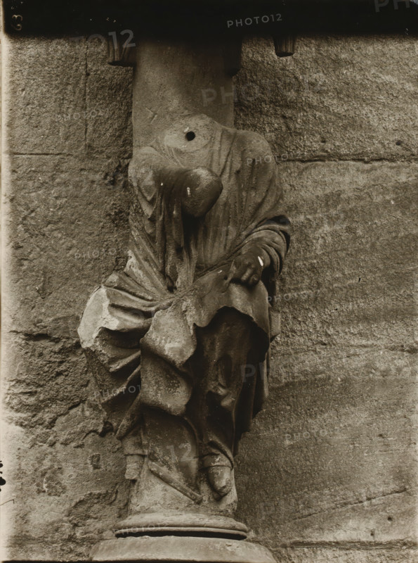 Detail of the Fürstenportal in the cathedral of Bamberg, Adamspforte, Johannes vN, album page, photograph, This photo is part of an album., number: '63', inscription: 'Kunsthist. Seminar, Marburg (Lahn), Photogr. Abteilung, Vervielfältigung, untersagt.', Bamberg, 1900 - c. 1920, photographer, Bavaria, paper, baryta paper, gelatin silver print, developing gelatin silver print, height 238 mm x width 180 mm, church (exterior), Bamberg Cathedral, A weathered stone sculpture of a draped figure mounted against a rough stone wall, showing folded and flowing fabric over a torso and limbs, with a rounded protrusion where a head might be and a circular hole in the upper chest area; the sculpture rests on a cylindrical stone plinth and displays chips, discoloration, and surface wear across the folds and edges, while the background consists of textured masonry blocks with visible joints and worn surfaces.