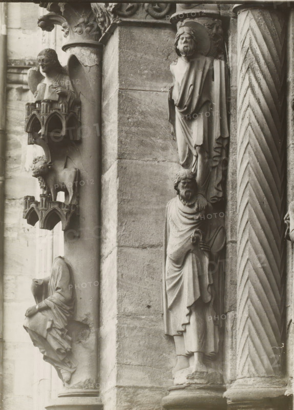 Adamspforte, Evangelisten u. Statuensäule li.lvN, Detail of the Fürstenportal in Bamberg Cathedral, album page, photograph, This photo is part of an album., inscription: 'Kunsthist. Seminar, Marburg (Lahn), Photogr. Abteilung, Vervielfältigung, untersagt.', number: '64', Bamberg, 1900 - c. 1920, photographer, Bavaria, paper, baryta paper, gelatin silver print, developing gelatin silver print, height 237 mm x width 176 mm, church (exterior), Bamberg Cathedral, A close view of carved stone figures and architectural details on a building façade, showing sculpted human figures in draped clothing with carved facial features and beards, a seated figure holding an object, a small animal figure standing on a decorative ledge with miniature arch motifs, ornate column capitals with floral and scroll patterns, a fluted column with diagonal chevron carving, and blocks of masonry with visible joints and weathering.