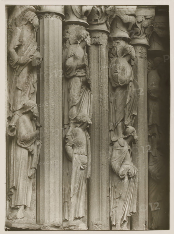 Detail of the Fürstenportal in Bamberg Cathedral, Evangelistensäule Stat. li. 2.3.4.vN, album page, photograph, This photo is part of an album., inscription: 'Kunsthist. Seminar, Marburg (Lahn), Photogr. Abteilung, Vervielfältigung, untersagt.', inscription: '67, Bamberg', Bamberg, 1900 - c. 1920, photographer, Bavaria, paper, baryta paper, gelatin silver print, developing gelatin silver print, height 236 mm x width 179 mm, church (exterior), Bamberg Cathedral, A close view of a stone architectural relief showing a row of tall fluted columns with carved human figures standing in recessed niches between them, each figure draped in flowing robes with visible folds and varying head coverings or hairstyles; some faces are in profile and others angled, with hands positioned near chests or holding drapery, and smaller figures or heads appear at shoulder or knee level of larger figures; the capitals above the columns feature carved foliage and decorative motifs, and the stone surface shows su...