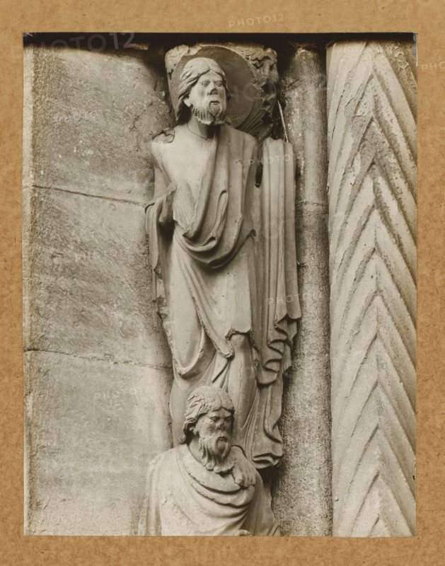 Detail of the Fürstenportal in Bamberg Cathedral, Evangelistensäule Apostel li.lvNW., album page, photograph, This photo is part of an album., number: '71', Bamberg, 1900 - c. 1920, photographer, Bavaria, paper, baryta paper, gelatin silver print, developed gelatin silver print, height 208 mm x width 162 mm, church (exterior), Bamberg Cathedral, A grayscale photograph shows two stone sculpted figures mounted on a textured wall, one positioned above the other; the upper figure wears draped robes and a halo, has a beard and long hair, and holds a fold of cloth at chest level, while the lower figure also has a beard and draped garments and appears with a turned head; to the right of the figures is a vertical carved column with a chevron pattern, and the surrounding stone surface shows weathered texture and horizontal joint lines, all framed by a mottled rectangular border.