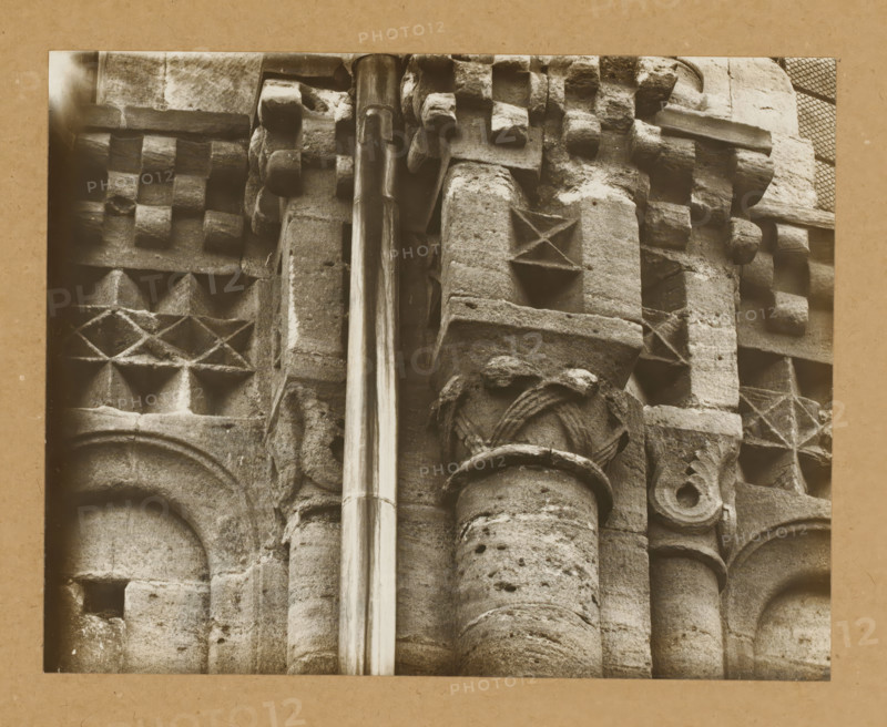 East Church, 1. Frieze 5., View of the sculpture of a frieze of Bamberg Cathedral, album page, photograph, This photo is part of an album., number: '7.', Bamberg, 1900 - c. 1920, photographer, Bavaria, paper, baryta paper, gelatin silver print, developed gelatin silver print, height 167 mm x width 211 mm, church (exterior), Bamberg Cathedral, A close view of aged stone architectural details showing carved blocks and columns with weathered surfaces, patterned geometric carvings including raised square and diamond shapes, rounded column shafts with fluted and pockmarked textures, a smooth vertical metal pipe running down the center, and fragments of ornamental molding and carved rosettes, all in a monochrome sepia tone with a plain border around the image.