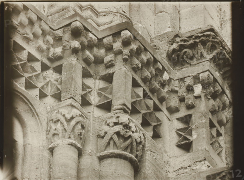 East Church, 1. Frieze 6., View of the sculpture of a frieze of Bamberg Cathedral, album page, photograph, This photo is part of an album., inscription: 'Kunsthist. Seminar, Marburg (Lahn), Photogr. Abteilung, Vervielfältigung, untersagt.', Bamberg, 1900 - c. 1920, photographer, Bavaria, paper, baryta paper, gelatin silver print, developed gelatin silver print, height 178 mm x width 235 mm, church (exterior), Bamberg Cathedral, A close-up view of a carved stone surface showing architectural details including rounded columns with smooth shafts and molded capitals, geometric star-like recessed patterns carved into square panels, rows of small rounded boss-like projections, and intricately carved foliage motifs on a corner capital; the stone has a weathered texture with visible tool marks and chips, and the image is monochrome with a soft vignette around the edges.