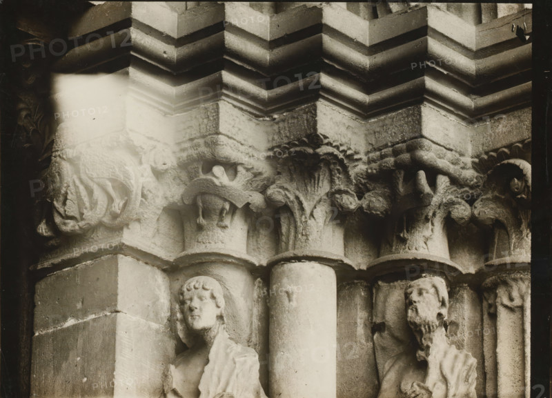 Detail of the Fürstenportal in Bamberg Cathedral, Fürstentor, Kapitelle r. über 1.2.v.NO., album page, photograph, This photo is part of an album., inscription: 'Bamberg, 83', inscription: 'Kunsthist. Seminar, Marburg (Lahn), Photogr. Abteilung, Vervielfältigung, untersagt.', Bamberg, 1900 - c. 1920, photographer, Bavaria, paper, baryta paper, gelatin silver print, developing gelatin silver print, height 177 mm x width 238 mm, church (exterior), Bamberg Cathedral, A close view of a stone architectural detail showing carved capitals and columns with layered mouldings above, featuring sculpted foliage motifs and animal-like forms, with two carved human figures set into vertical niches beside the columns, one with a youthful face and short curly hair and the other with a bearded face and draped clothing, all rendered in weathered pale stone with areas of shadow and darker grime and a bright washed area near the upper left.