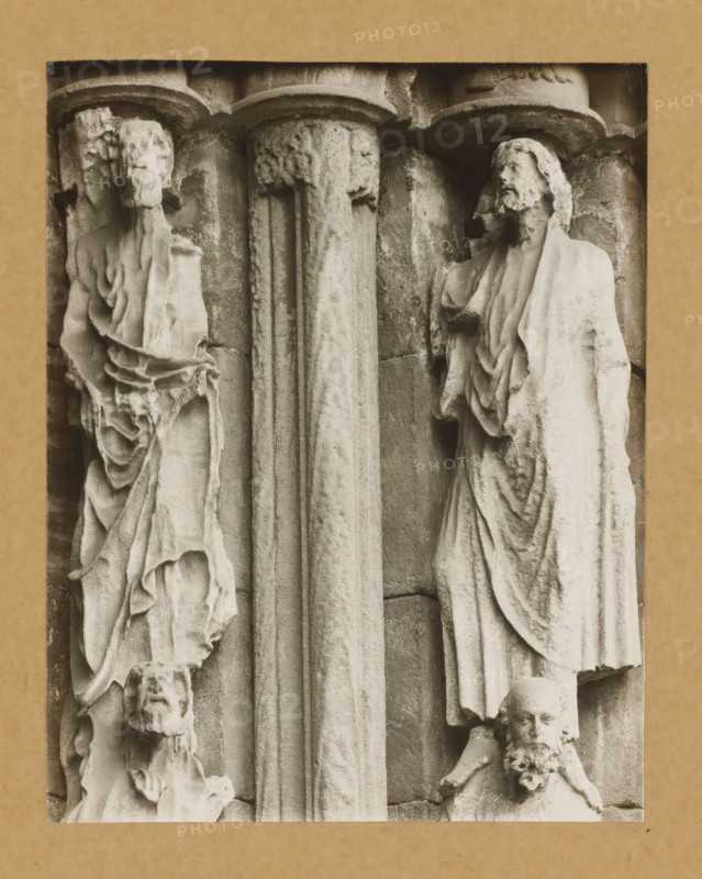 Fürstentor, Ap. r.3.4.v.NO., Detail of the Princely Portal in Bamberg Cathedral, album page, photograph, This photo is part of an album., number: '85', Bamberg, 1900 - c. 1920, photographer, Bavaria, paper, baryta paper, gelatin silver print, developed gelatin silver print, height 208 mm x width 164 mm, church (exterior), Bamberg Cathedral, A close-up view of a stone relief framed by a rectangular border, showing two vertical fluted columns in the center with carved capitals, flanked by carved human figures in draped garments; on the left a weathered figure with a long draped robe and an eroded face, and below it a smaller carved head with a beard, while on the right a standing robed figure with a bearded head and detailed folds in the fabric, and below that another carved bearded head with a distinct floral-like beard detail; the stone surface shows texture, wear, and subtle shadows emphasizing the carved folds and architectural elements.