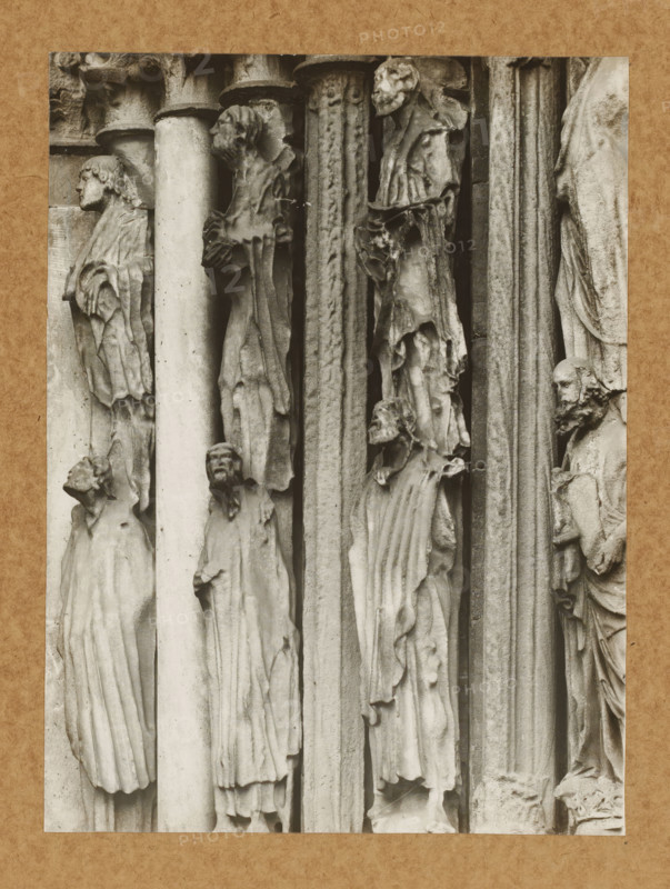 Detail of the Princely Portal in Bamberg Cathedral, Princely Gate Statue no. 1.2.3. vN, album page, photograph, This photo is part of an album., number: '91', Bamberg, 1900 - c. 1920, photographer, Bavaria, paper, baryta paper, gelatin silver print, gelatin silver developing print, height 211 mm x width 158 mm, church (exterior), Bamberg Cathedral, A sepia-toned photograph of a carved stone façade showing a vertical arrangement of elongated human figures in robes positioned between columns and narrow pilasters; the figures display varied head poses and facial features with some heads turned in profile and others facing forward, and the drapery of their garments forms deep vertical folds and textured surfaces; the stone shows weathering and roughness with cracks and chips visible along edges, and narrow shadowed recesses separate the figures and architectural elements; a textured paper border frames the image.