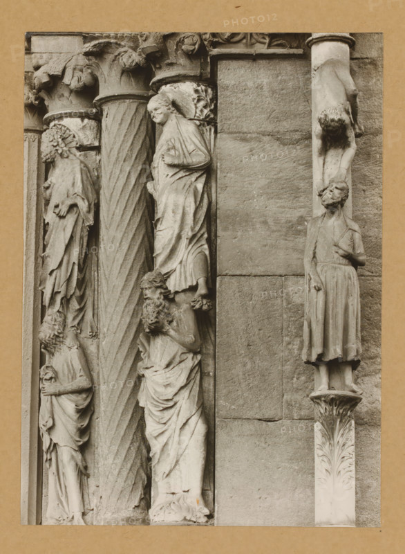 Detail of the Princely Portal in Bamberg Cathedral, Princely Gate Statues of the 5th and 6th century of Judas Column, album page, photograph, This photo is part of an album., number: '93', Bamberg, 1900 - c. 1920, photographer, Bavaria, paper, baryta paper, gelatin silver print, height 221 mm x width 160 mm, church (exterior), Bamberg Cathedral, A vertical stone relief showing carved figures and decorative columns set into a wall panel, with several robed human figures in various poses and expressions carved in a weathered stone texture, a twisted fluted column between groups of figures, ornate capitals with acanthus-like carvings, a figure suspended upside down attached to a narrow column, and a standing figure on a carved plinth, all rendered in a monochrome stone tone against a plain masonry background.