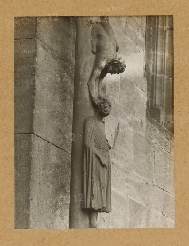 Detail of the Princely Portal in Bamberg Cathedral, Princely Gate Judas Column, the figure, album page, photograph, This photo is part of an album., number: '94.', Bamberg, 1900 - c. 1920, photographer, Bavaria, paper, baryta paper, gelatin silver print, height 226 mm x width 162 mm, church (exterior), Bamberg Cathedral, A grayscale photograph shows a carved stone figure attached to a vertical column against a textured stone wall; the figure is draped in a long garment with visible folds and stands upright while another smaller carved figure appears above, bending forward with one arm extended downward toward the head of the lower figure; the upper figure has stylized hair and a rounded form, and the lower figure's facial features and drapery are visible; the stone surfaces show weathering, grain, and subtle discoloration, and the image is bordered by a narrow lighter frame and a wider textured mat.