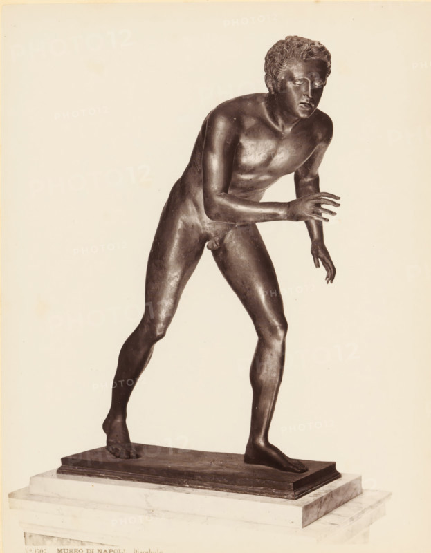 Bronze sculpture of an athlete, Museo di Napoli Discobolo, album page, photograph, This photo is part of an album., number: '1505', workshop stamp: 'Giorgio Sommer, Largo Vittoria, Napoli', photographer: Giorgio Sommer, Naples, 1874 - in or before 1879, paper, albumen print, height 251 mm x width 199 mm, sculpture (bronze, arts), athletics, gymnastics (sportsman, player, etc.), A dark-toned bronze statue of a nude young male figure is shown standing on a rectangular base placed on a marble plinth; the figure leans forward with one leg stepped ahead and the other trailing, one arm extended forward with fingers splayed and the other arm lowered and slightly bent, the head turned slightly to the side with curly hair and a focused facial expression, the surface of the bronze shows subtle highlights and patina, and the background is a plain light color.