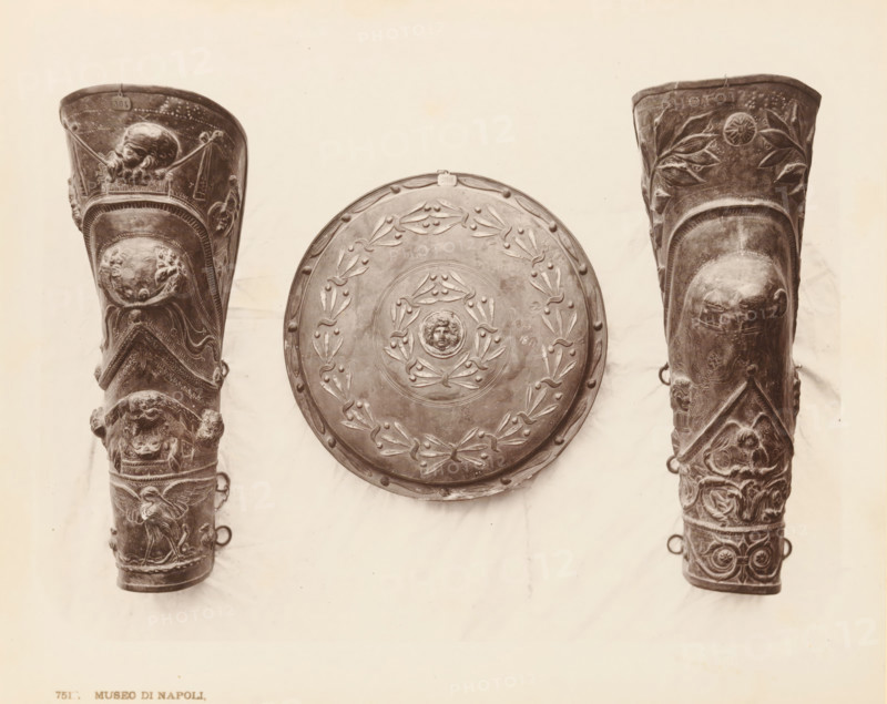 Decorated objects, Museo di Napoli Saffo, album page, photograph, This photo is part of an album., workshop stamp: 'Giorgio Sommer, Largo Vittoria, Napoli', number: '7517', photographer: Giorgio Sommer, Naples, 1874 - in or before 1879, paper, albumen print, height 202 mm x width 254 mm, military clothing and other equipment (uniforms, cap, armor, helmet, etc.), A centered round shield with raised decorative motifs and a small sculpted face at its center hangs between two tall, elaborately carved cylindrical objects; the shield shows a wreath-like pattern and small round studs along its rim. The left cylindrical object features multiple relief scenes including a bearded head near its top, ornate bands and panels with animal and human figures, and loops protruding from its side. The right cylindrical object mirrors the left in shape and also displays relief carvings including vegetal motifs near the top, a rounded protuberance with facial features set into a recessed panel, intertwined ...