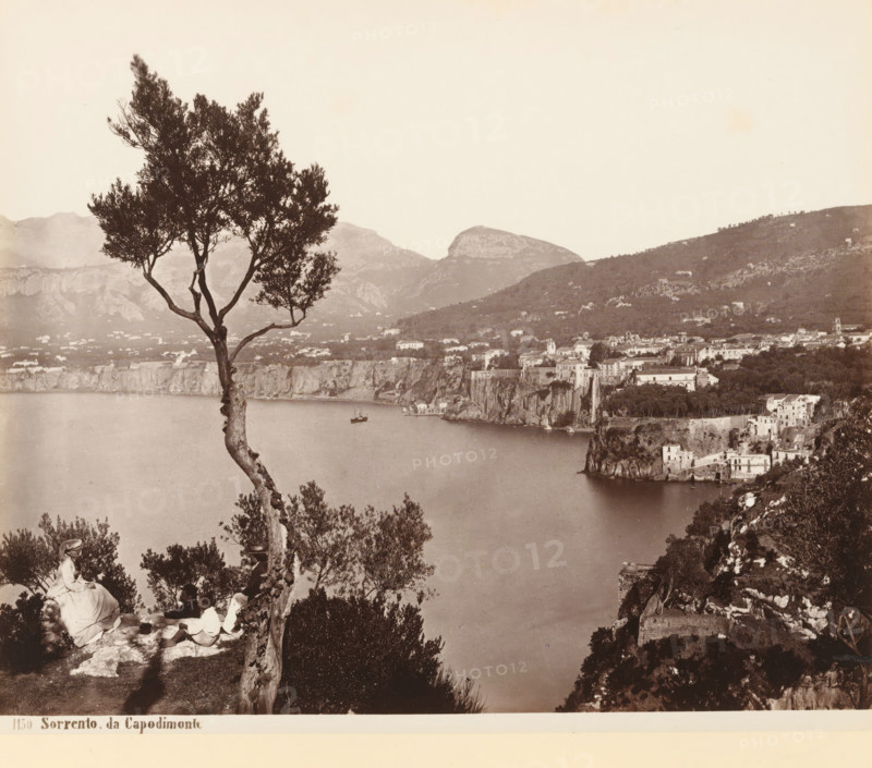 View of a body of water near Capodimonte, Sorento da Capodimonte, album page, photograph, This photo is part of an album., number: '1150', workshop stamp: 'Giorgio Sommer, Largo Vittoria, Napoli', photographer: Giorgio Sommer, Capodimonte, 1874 - in or before 1879, Naples, paper, albumen print, height 204 mm x width 243 mm, trees, landscapes with waters, waterscapes, seascapes (in the temperate zone), mountains, A coastal landscape seen from a high vantage point with a tall, slender tree in the foreground beside rocky ground where seated figures rest; below is a calm body of water with a small boat and a distant shoreline of cliffs and buildings hugging the coast, while terraced and clustered structures extend up a hillside and wooded slopes rise behind them beneath a pale sky.