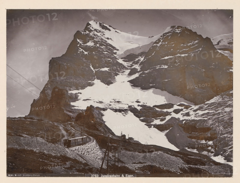 Jungfraubahn & Eiger, Jungfraubahn railway near the Eiger, photograph, This photo is part of a cover., inscription: 'Gebr. Wehrli, Kilchberg Zürich 3783 Jungfraubahn & Eiger.', Kleine Scheidegg, publisher: Gebrüder Wehrli, Kilchberg, 1912 - 1920, photographer, paper, pasteboard, baryta paper, daylight collodion silver print, height 163 mm x width 219 mm, railway, train, mountains, Alpen, Bernese Alps, Jungfrau, Eiger, A large snow-covered mountain with steep rocky faces fills the image, showing patches of snow and exposed dark rock on its slopes and summit. A narrow railway track winds up the lower slope, with a rail car positioned on the track and utility poles with overhead lines running alongside. The foreground shows rocky ground and a small path leading toward the track, with a few small structures near the base of the mountain. The sky above the peak is overcast and the image has an aged, sepia-toned appearance.