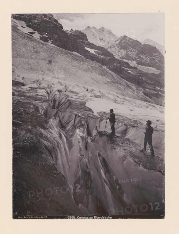 Crevasse am Eigergletscher, Two men at a crevasse in the Eiger Glacier, photograph, This photo is part of a cover., inscription: 'Gebr. Wehrli, Kilchberg, Zürich. 3665. Crevasse am Eigergletscher', Eiger, publisher: Gebrüder Wehrli, 1897 - 1920, photographer, Alps, Bernese Alps, paper, pasteboard, baryta paper, daylight collodion silver print, height 220 mm x width 163 mm, ravine, chasm, abyss, canyon, glacier, mountain climbing, mountaineering, A black and white photograph of a mountainous icy landscape showing a large glacier with deep crevasses and smooth, striated ice surfaces extending up a steep slope toward rocky, snow-dusted peaks in the background; two men in dark clothing and hats stand on the glacier near the edge of a deep fissure, one holding a long pole and the other holding a rope, both facing the crevasse; jagged rock faces and patches of snow appear on the higher slopes behind them, and the sky above is pale with scattered clouds.