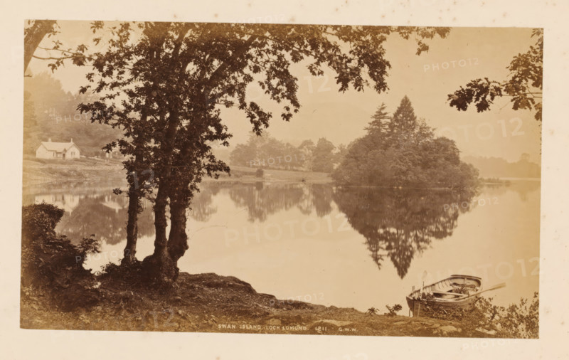 View of Swan Island in Loch Lomond, Swan Island, Loch Lomond, photograph, number: '1211, GWW', photographer: George Washington Wilson, Scotland, 1865 - 1880, paper, albumen print, height 116 mm x width 192 mm, island, landscapes with waters, waterscapes, seascapes (in the temperate zone), A sepia-toned lakeside scene with a calm body of water reflecting trees and shoreline. In the foreground left, a large tree with multiple trunks and overhanging branches frames the view, its roots and surrounding ground visible. Near the right foreground, a small wooden rowboat rests at the water’s edge with oars and interior details visible. Across the lake, a densely treed island rises with a mix of rounded foliage and taller coniferous shapes, its reflection mirrored in the water. On the far left shore, a light-colored cottage with a pitched roof sits among grassy banks and scattered trees. The sky appears pale and hazy above the distant tree line.