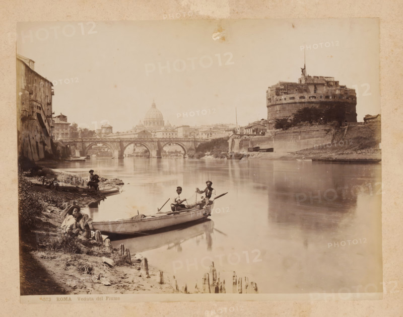 View of the Tiber River in Rome, album page, photograph, This photo is part of an album., inscription: '573 ROMA Veduta del Fiume', Rome, 1870 - c. 1895, photographer, paper, pasteboard, albumen print, height 200 mm x width 258 mm, prospect of city, town panorama, silhouette of city, canals, waters (in city), Tiber, A riverside scene shows calm water reflecting a stone bridge with multiple arches and buildings lining the far bank, including a large cylindrical fortress with battlements and a statue on its roof; nearer the left bank there are old multi-story buildings with shuttered windows and a sloped embankment leading to the water where small boats are moored and wooden stakes protrude from the muddy shore; several men sit and stand in and beside the boats, some holding oars or fishing nets, wearing hats and simple clothing; distant domed architecture rises behind the bridge, and trees and low walls dot the riverbanks under a pale sky.