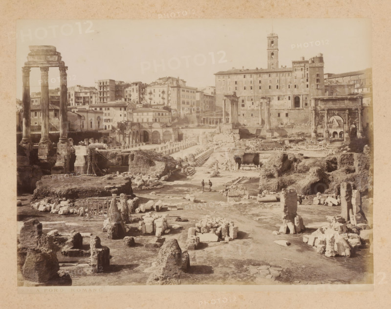 View of the Roman Forum in Rome, album page, photograph, This photo is part of an album., inscription: '292 FORO ROMANO', Rome, 1870 - c. 1895, photographer, paper, pasteboard, albumen print, height 198 mm x width 257 mm, ruin of a building, architecture, Roman Forum, A wide sepia photograph of an archaeological site showing scattered stone ruins and fragmented columns across uneven ground, with three tall standing columns with carved capitals on the left and a large archway structure with decorative details on the right, a long multi-story building with many windows and a tower rising behind the central area, rows of arches and stone stairways amid the ruins, rubble piles and stone blocks arranged in the foreground, and two small human figures walking along a pathway near the center of the scene.