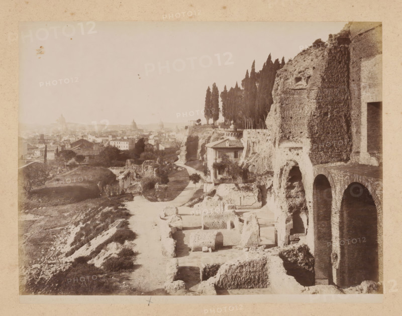 View of Rome from the Palatine Hill, album page, photograph, This photo is part of an album., inscription: '37. VEDUTA DI ROMA DAL PALATINO', Rome, 1870 - c. 1895, photographer, paper, pasteboard, albumen print, height 200 mm x width 258 mm, prospect of city, town panorama, silhouette of city, A sepia-toned view of an ancient stone ruin on a hillside with large arched openings and weathered brickwork along the right edge, terraced stone blocks and carved niches visible in the foreground, a dirt path winding past scattered rectangular stone remains toward a small building with a tiled roof set into the cliff, a line of tall, narrow cypress trees rising behind the cliff edge, cultivated plots and trees on lower slopes, a distant urban skyline of low buildings and domed rooftops across the horizon, and a pale sky filling the upper portion of the image.