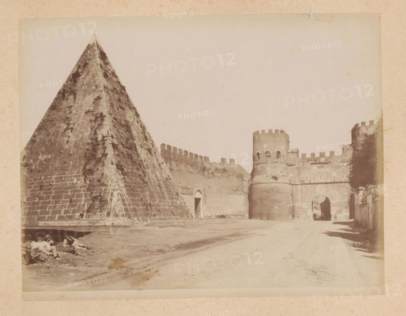 View of the Pyramid of Cestius and the Porta San Paolo in Rome, album page, photograph, This photo is part of an album., inscription: '137 PIRAMIDE DI GAIO CESTIO CON PORTA S. PAOLO', Rome, 1870 - c. 1895, photographer, paper, pasteboard, albumen print, height 201 mm x width 259 mm, city-gate, pyramid (marking grave), Porta San Paolo, Pyramid of Cestius, A large stone pyramid with weathered, patterned blocks fills the left side, showing a pointed top and dark streaks on its surface, while to the right a fortified stone wall with crenellations stretches toward cylindrical towers with rounded battlements and arched openings, one tower featuring small window-like apertures and another forming an arched gateway leading into a shadowed passage; a dirt road runs in front of the structures with faint wheel or track marks, a shadowed tree or foliage appears near the rightmost tower, and a group of people recline on the ground in the shaded area at the pyramid’s base.