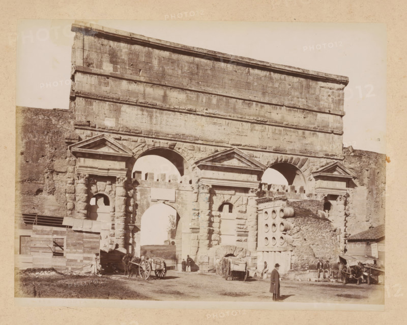 View of the Porta Maggiore in Rome, album page, photograph, This photo is part of an album., inscription: '448 PORTA MAGGIORE', Rome, 1870 - c. 1895, photographer, paper, pasteboard, albumen print, height 200 mm x width 259 mm, city-gate, Porta Maggiore, A sepia photograph of a large stone archway and façade showing weathered masonry, carved pediments above arched openings, and stacked horizontal stone courses, with rough textured walls on either side; in front of the structure there are wooden carts and horses, a few people standing and sitting near the entrance, a man in a long coat and hat walking in the foreground, a temporary wooden building with corrugated roofing to the left, and scattered rubble and architectural fragments on the ground.