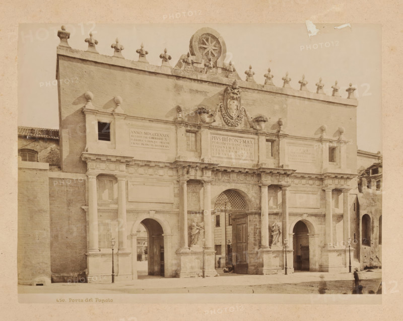 View of the Porta del Popolo in Rome, album page, photograph, This photo is part of an album., inscription: '450. Porta del Popolo', Rome, 1870 - c. 1895, photographer, paper, pasteboard, albumen print, height 199 mm x width 260 mm, city-gate, Porta del Popolo, A sepia-toned photograph of a large stone gateway with classical architectural elements, featuring tall columns and arched openings at ground level, decorative statues positioned in niches, ornate carvings and sculpted reliefs above the central entrance, a row of small sculpted finials along the top edge of the facade, a circular decorative element with a star-like motif near the roofline, lamp posts on the pavement in front of the structure, and a blurred figure walking in the foreground.