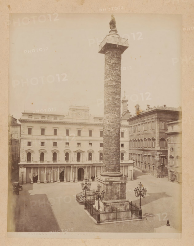 View of the column of Marcus Aurelius on the Piazza Colonna in Rome, album page, photograph, This photo is part of an album., inscription: '.32 COLONNA DI MARCO AURELIO', Rome, 1870 - c. 1895, photographer, paper, pasteboard, albumen print, height 259 mm x width 200 mm, triumphal column with helical friezes, Column of Marcus Aurelius, A tall, intricately carved stone column rises from a square base surrounded by a low iron fence, topped by a statue on a platform with railings; the column stands in a paved open square with ornate street lamps nearby and a few small decorative bollards, flanked by multi-story classical buildings with rows of windows, arched ground-level openings and columned porticoes, and a building facade with a clock feature above its upper windows; the scene is monochrome and appears framed by a light border.