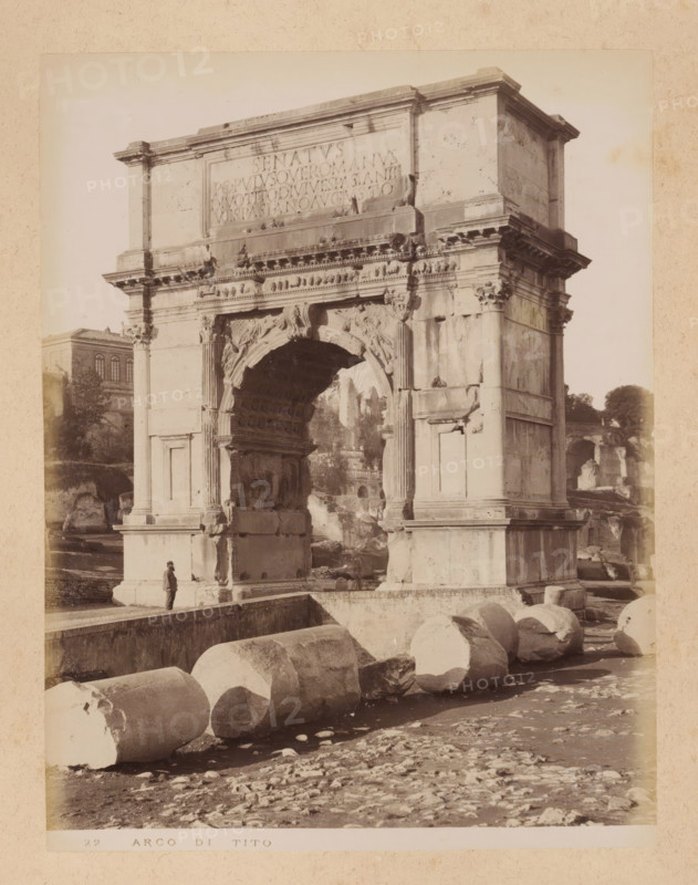 View of the Arch of Titus in the Roman Forum in Rome, album page, photograph, This photo is part of an album., inscription: '22 ARCO DI TITO', Rome, 1870 - c. 1895, photographer, paper, pasteboard, albumen print, height 258 mm x width 199 mm, triumphal arch, Arch of Titus, A large stone archway with decorative carvings and fluted columns dominates the scene, showing weathered surfaces and worn ornamentation, while a person stands near its base providing scale. Broken cylindrical stone drums lie scattered in the foreground on uneven ground. Behind and around the arch are ruined masonry and fragments of structures, with a building partially visible in the midground to the left and trees in the background. The image has a sepia tone and shows the monument mounted on a raised platform with steps and ledges.