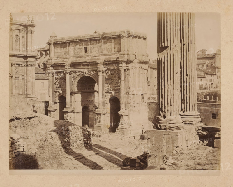 View of the Arch of Septimius Severus on the Roman Forum in Rome, album page, photograph, This photo is part of an album., inscription: '18 ARCO DI SEPTIMIO-SEVERO', Rome, 1870 - c. 1895, photographer, paper, pasteboard, albumen print, height 199 mm x width 258 mm, triumphal arch, Arch of Septimius Severus, A sepia-toned photograph of a large ancient stone arch and surrounding ruins with carved reliefs and architectural details, flanked on the right by a tall fluted column standing on a multi-tiered base and on the left by part of a classical building façade with decorative pilasters and sculptural elements; the arch shows multiple openings and ornate carvings above and around the central arched passage, with worn stone surfaces and broken fragments visible; in the foreground uneven ground, rubble, and collapsed masonry create rough paths and shadows leading toward the structure, and a distant cluster of tiled-roof buildings is visible beyond the column.