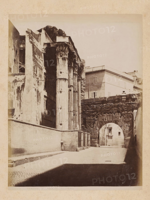 View of the Temple of Mars Ultor at the Forum of Augustus in Rome, album page, photograph, This photo is part of an album., inscription: '503 TEMPLO DU MARTE ULTORE', Rome, 1870 - c. 1895, photographer, paper, pasteboard, albumen print, height 260 mm x width 199 mm, ruin of a building, architecture, (story of) Mars (Ares), temple, shrine, Roman religion, Forum of Augustus, Temple of Mars Ultor, A sepia photograph shows a narrow street flanked by high walls and ancient stone structures, with tall, fluted columns topped by ornate capitals visible on the left and an arched stone passageway spanning the street further back on the right; buildings with shuttered windows sit above the arch, and a street lamp is mounted on the wall near the arch; the ground is paved and a deep shadow falls across the foreground of the scene.