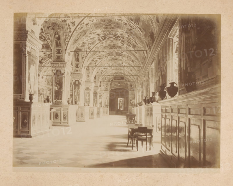 View of the Sistine Chapel in the Apostolic Palace in Vatican City, album page, photograph, This photo is part of an album., inscription: '385 SALA DELLA ... VATICANO', Sistine Chapel (Vatican), 1870 - c. 1895, photographer, Vatican City, Apostolic Palace, paper, pasteboard, albumen print, height 200 mm x width 260 mm, parts of church interior: chapel, A long ornate interior with a vaulted ceiling covered in painted panels and decorative motifs, tall pillars and arches lining a central corridor, framed paintings and sculpted figures set into wall panels, a row of dark vases displayed on a continuous ledge along one side, wooden cabinets or paneling beneath the ledge, a small wooden table and chairs placed on the tiled floor, checkerboard floor pattern visible in the foreground, soft light casting shadows across the floor and furnishings, and a distant doorway or window at the far end of the corridor.