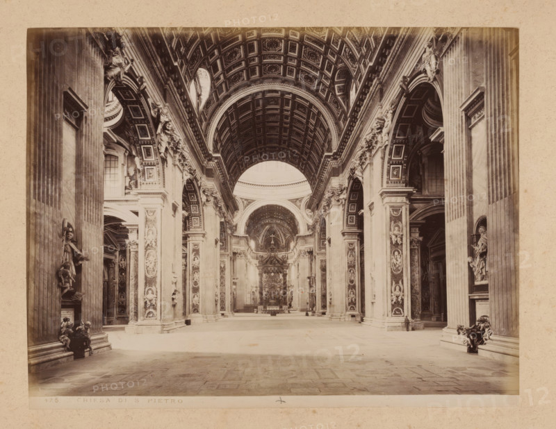 Interior of St. Peter's Basilica in Vatican City, album page, photograph, This photo is part of an album., inscription: '175 CHIESA DI S PIETRO', St. Peter's Basilica, 1870 - c. 1895, photographer, Vatican City, paper, pasteboard, albumen print, height 200 mm x width 258 mm, interior of church, A wide interior view of a grand church nave with a coffered barrel-vaulted ceiling and large arched openings along the sides, flanked by tall fluted columns and richly decorated pilasters with sculpted reliefs and statues set into niches, ornamental cornices and carved capitals; the central aisle leads toward an ornate baldachin and altar area visible in the distance, with layered arches and detailed ornamentation above, while figures of sculpted saints and decorative angels occupy many of the vertical surfaces and pedestals, and the stone floor shows a patterned surface extending to the foreground.