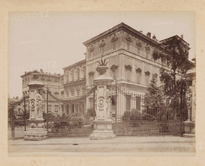 View of the Palazzo Barberini in Rome, album page, photograph, This photo is part of an album., inscription: '60 IL CAMPIDOGLIO', Rome, 1870 - c. 1895, photographer, paper, pasteboard, albumen print, height 199 mm x width 258 mm, palace, Palazzo Barberini, A sepia-toned photograph showing the façade of a large ornate building behind an iron fence, with a central corner section featuring shuttered windows and decorative window pediments, fluted pilasters, and a pronounced cornice; in front of the fence are two tall stone pillars topped with decorative urns and sculptural reliefs, and a curved wrought-iron gate with intricate scrollwork; behind the fence are planted shrubs and taller trees partially obscuring the right side of the building, and the street and sidewalk run across the foreground.