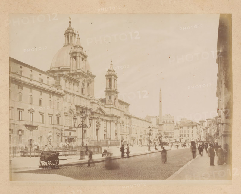 View of the Piazza Navona in Rome, album page, photograph, This photo is part of an album., inscription: '423 PIAZZA NAVONA', Rome, 1870 - c. 1895, photographer, paper, pasteboard, albumen print, height 200 mm x width 259 mm, square, circus, Piazza Navona, A wide sepia photograph showing a large ornate stone church with a prominent dome and flanking towers, detailed facades, and decorative cornices, a broad cobblestone street in front, ornate street lamps and a fenced plaza area with sculptures and fountains, groups of people standing and walking along the sidewalk and near the plaza, a cart on the street, rows of multi-story buildings receding into the distance, and an obelisk visible in the background under a pale sky.