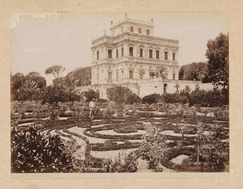 View of the Villa Doria Pamphilj in Rome, album page, photograph, This photo is part of an album., inscription: '615 G - Villa Pamfili', Rome, 1870 - c. 1895, photographer, paper, pasteboard, albumen print, height 198 mm x width 259 mm, villa, park, Janiculum, Villa Doria Pamphili, A sepia-toned photograph shows a formal garden with low clipped hedges arranged in concentric and curving patterns in the foreground, interspersed with gravel pathways and potted plants on raised bases. Behind the patterned beds, taller ornamental shrubs and small trees form a dense boundary, with a person wearing light clothing and a hat standing among the plants near a central path. A multi-story classical villa with ornate façade details, rows of windows, sculptural reliefs, decorative cornices, and a terrace with balustrade rises behind the garden. Mature trees, including a tall umbrella-shaped tree, frame the villa on either side, and potted plants sit on the villa's terrace and stair landing. The image...