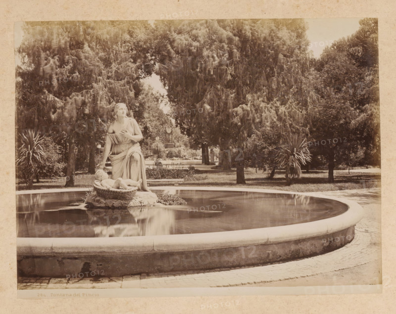 View of a fountain on the Pincio in Rome, album page, photograph, This photo is part of an album., inscription: '284. Fontana del Pincio', Rome, 1870 - c. 1895, photographer, paper, pasteboard, albumen print, height 195 mm x width 257 mm, ornamental fountain, Pincio, A sepia-toned photograph shows a circular stone fountain with still water reflecting a central sculptural group; the sculpture depicts a robed female figure seated on a decorative base with one hand near her chest and the other touching a reclining child figure within a carved basin, and the figures rest on ornate stonework at the fountain center; the fountain rim curves around the foreground and sits on a paved area; behind the fountain a formal garden area is visible with trimmed lawns, tall trees with dense drooping foliage, and clusters of spiky-leaved plants, while further back a stone columnar structure and garden benches appear partially obscured by the trees.