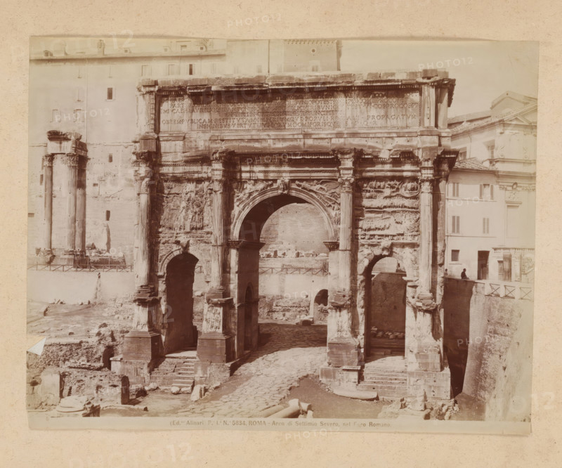 View of the Arch of Septimius Severus at the Roman Forum in Rome, album page, photograph, This photo is part of an album., inscription: '(Ed.ne Alinari) Pa Ia No 5834. ROMA - Arco di Settimio Severo, nel Foro Romano.', photographer: Fratelli Alinari (possibly), Rome, 1870 - c. 1895, paper, pasteboard, albumen print, height 192 mm x width 247 mm, triumphal arch, Arch of Septimius Severus, A large stone triumphal arch with three arched openings fills the foreground, showing worn columns, carved reliefs, and decorative moldings; the central arch is the tallest and flanked by paired columns with sculpted details, while the lower sections reveal stepped bases and eroded stonework. Behind and beside the arch are multi story buildings with plain facades and shuttered windows, and a tall isolated pair of standing columns is visible to the left. The ground in front of and beneath the arch consists of uneven cobbles, scattered stone fragments, and exposed foundations. A low railing runs along th...