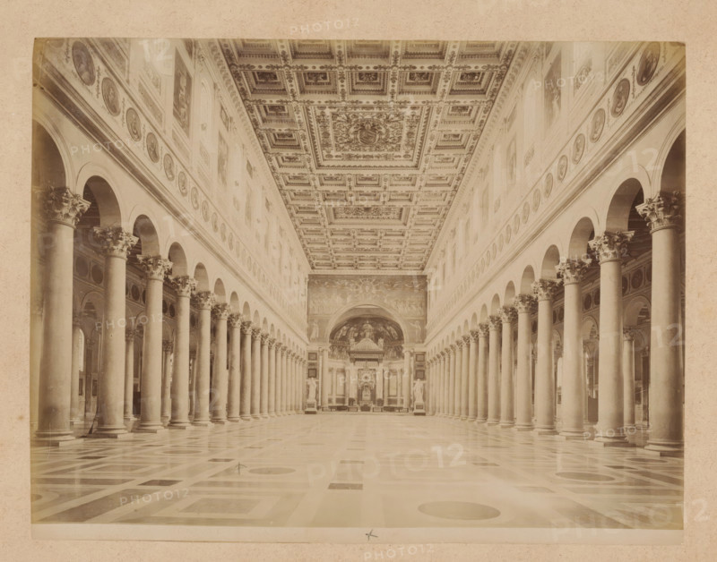 Interior of St. Paul Outside the Walls in Rome, album page, photograph, This photo is part of an album., inscription: '... FIORE LE MURA', St. Paul Outside the Walls, 1870 - c. 1895, photographer, Rome, Saint Paul Outside the Walls, paper, pasteboard, albumen print, height 199 mm x width 258 mm, interior of church, A wide interior view of a long, columned hall with two parallel rows of tall stone columns topped by ornate capitals and connected by rounded arches, a patterned floor with geometric shapes, a richly decorated coffered ceiling with square panels and an elaborate central design, circular medallion-like decorations along the upper walls, large arched openings above the columns, sculpted statues placed near the far end, and a decorated apse area with painted scenes and architectural ornamentation.
