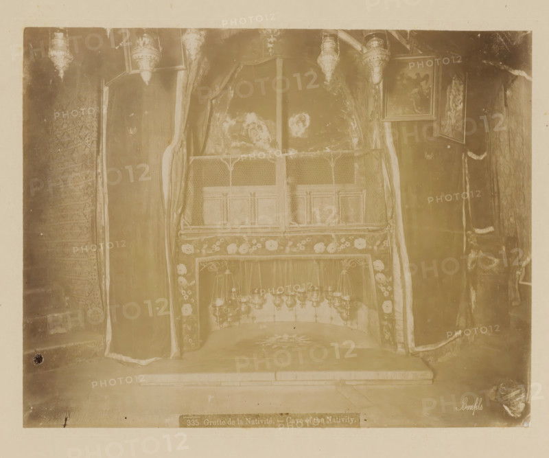 Interior of the Church of the Nativity in Bethlehem, Grotte de la Nativité, Cave of the Nativity, album page, photograph, This photo is part of an album., number: '335', photographer: Félix Bonfils, Bethlehem (Israel), 1867 - 1885, Palestine, paper, albumen print, height 218 mm x width 277 mm, interior of church, Church of the Nativity, A faded photograph of an ornate alcove framed by draped curtains, with decorative panels and patterned fabric visible around the opening, a low stone step leading into the recessed space, a patterned rug or floor covering inside the alcove, a row of hanging lamps or chandeliers suspended within the recess, carved or paneled surfaces above the opening, and framed paintings hanging on the wall near the top edge of the alcove.